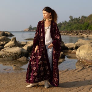 May include: A long, burgundy velvet robe with a floral pattern and dark purple trim. The robe is worn over a white top and blue jeans. The background shows a beach with rocks and the ocean.