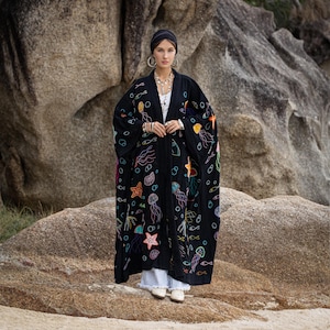 Op de afbeelding: Een zwarte kimono-achtige robe met kleurrijke geborduurde zeedieren, waaronder kwallen, zeesterren en vissen. De robe wordt gedragen over een witte top en een lichtblauwe broek. De achtergrond toont grote rotsen.
