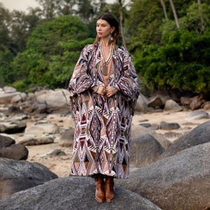 May include: A long, patterned kimono in shades of brown, white, and burgundy, worn over a neutral-colored top and paired with brown boots. The kimono features a geometric design. The person is standing on a large rock near a beach.