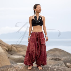 May include: A woman wearing a black crop top and maroon harem pants with pockets. The pants have a patchwork design and a drawstring waist. She is also wearing jewelry, including a necklace and bracelets. The background is a beach scene with rocks and the ocean.