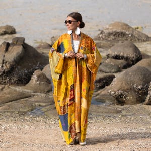 Puede incluir: Un kimono largo y fluido de color amarillo con un diseño de patchwork. El kimono presenta varios patrones y colores, incluyendo azul, naranja y marrón. La prenda se lleva al aire libre, con una playa y rocas en el fondo.