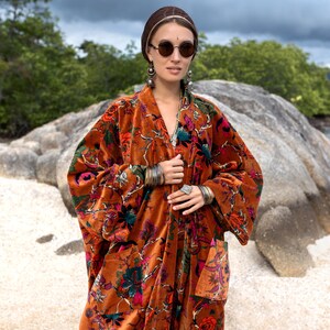 Close-up portrait of the Amber Flora Harper Jade Velvet Kimono Robe, photographed outdoors with rocky textures and green trees behind. The richly patterned velvet features jewel-toned florals blooming across a warm sienna base, complemented by oversized sleeves and patch pockets. Styled with silver bangles, earrings, and a head wrap, this boho plus size robe embodies artisan craftsmanship and autumnal elegance, perfect for festivals, parties, and everyday luxe comfort.