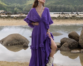 May include: A woman wearing a long, purple, flowy dress with a ruffled hem and a deep V-neckline. She is standing on a beach with rocks and water in the background.