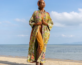 May include: A woman wearing a long, green, yellow, and red patterned kimono with a brown leather belt. She is standing on a sandy beach with the ocean in the background.