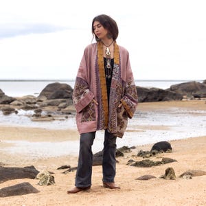 May include: A long-sleeved, open-front kimono jacket featuring a dusty rose base color with intricate, multi-colored patterned panels in shades of gold, brown, and deep purple. The garment includes two front pockets and a mustard yellow trim.