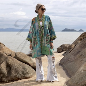 May include: A patchwork kimono in shades of teal, green, and floral patterns. The kimono has a tie closure and pockets. The model is wearing distressed white jeans and sunglasses. The background features a seascape with rocks and water.