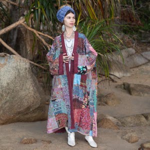 Puede incluir: Una bata kimono de patchwork en una variedad de colores, incluyendo rojo, azul y rosa. La bata tiene mangas largas y un ribete burdeos. La modelo lleva un pañuelo azul en la cabeza y zapatos blancos. La bata está hecha de cuadrados de tela.