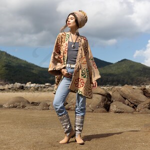 May include: A woman wearing a beige kimono with a floral pattern, blue jeans, and brown boots. She is standing on a beach with rocks and sand in the background.