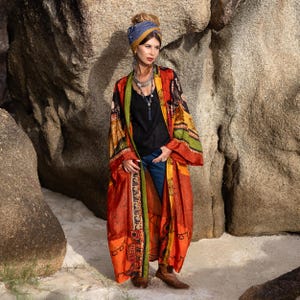 Könnte beinhalten: Ein langer, fließender Kimono in Rot-, Orange- und Gelbtönen mit aufwendigen Mustern. Der Kimono hat einen schwarzen Rand mit grünen Akzenten und ein detailliertes Design. Er wird über einem schwarzen Oberteil und blauen Jeans getragen, was einen Bohemian-Stil erzeugt.