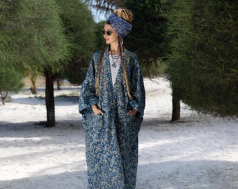 May include: A woman wearing a blue and white patterned long robe with a blue head scarf and sunglasses. She is standing in front of a white sandy beach with green trees in the background.