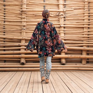 Back view of the Mei Velvet Kimono Duster in Nocturne Flora by ZinniaPearl. The model walks on a wooden deck framed by natural bamboo textures, showing the jacket’s oversized flow and dramatic sleeves. The black velvet canvas glows with detailed florals in ruby, saffron, and jade tones, symbolizing bohemian artistry and handmade heritage. A perfect visual of movement, grace, and slow fashion luxury.