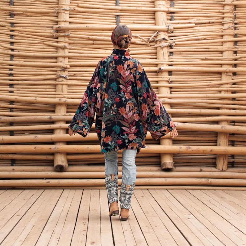 Back view of the Mei Velvet Kimono Duster in Nocturne Flora by ZinniaPearl. The model walks on a wooden deck framed by natural bamboo textures, showing the jacket’s oversized flow and dramatic sleeves. The black velvet canvas glows with detailed florals in ruby, saffron, and jade tones, symbolizing bohemian artistry and handmade heritage. A perfect visual of movement, grace, and slow fashion luxury.