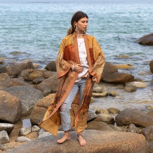 May include: A long, patterned kimono in gold, orange, and brown hues. The garment is worn over a white top and light blue jeans. The image is set on a rocky beach with the sea in the background. The kimono's design includes detailed patterns.