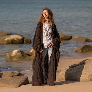 Front view of the VEDA Patchwork Duster in Cocoa Earth. A floor-sweeping, oversized kimono coat made from heavy stone-washed cotton canvas. The model holds the lapels, showing the open-front design, deep patch pockets, and the rich, chocolate brown color. The raw-style stitching creates a visible grid pattern, emphasizing the wabi-sabi artisan aesthetic.
