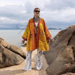 May include: A yellow, orange, and red patterned kimono-style jacket with long sleeves and a tie closure. The jacket features a patchwork design and is worn with light-wash, ripped jeans. The background includes a beach and ocean.