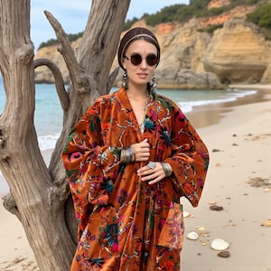 Close-up portrait of model in the Amber Flora Harper Jade Kimono Robe by ZinniaPearl. The velvet duster displays richly textured floral embroidery with jewel-toned botanicals on a burnt orange base. Styled with silver jewelry, sunglasses, and natural hair wrap, the look radiates bohemian goddess energy. The handcrafted velvet robe features oversized sleeves and deep pockets, designed for inclusive sizing from M to 4X, perfect as a fall statement coat or holiday gift.