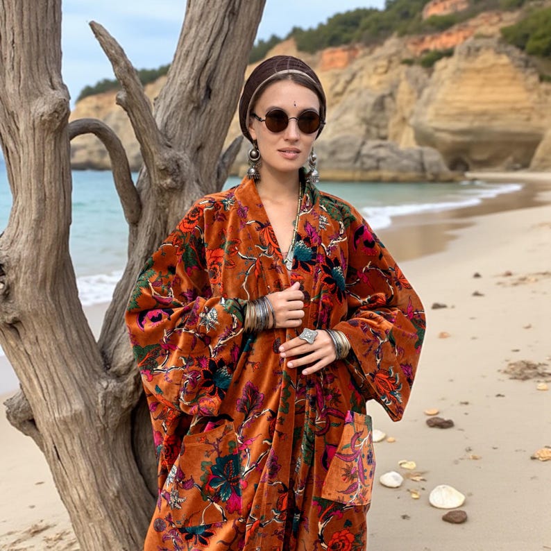 Close-up portrait of model in the Amber Flora Harper Jade Kimono Robe by ZinniaPearl. The velvet duster displays richly textured floral embroidery with jewel-toned botanicals on a burnt orange base. Styled with silver jewelry, sunglasses, and natural hair wrap, the look radiates bohemian goddess energy. The handcrafted velvet robe features oversized sleeves and deep pockets, designed for inclusive sizing from M to 4X, perfect as a fall statement coat or holiday gift.