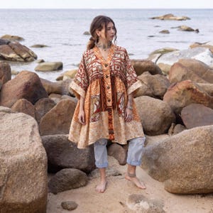 Op de afbeelding: Een ruimvallende tuniek met V-hals voorzien van een patchwork van patronen in warme tinten oranje, bruin en crème. Het kledingstuk heeft wijde mouwen tot op de elleboog, een knoopsluiting aan de voorzijde en een uitlopende zoom, gecombineerd met een lichtblauwe spijkerbroek.