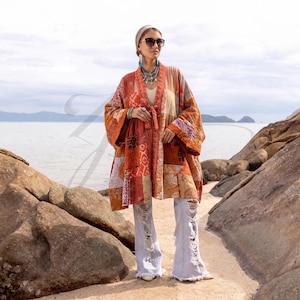 Op de afbeelding: Een kimono-jas met lange mouwen en een open voorkant, voorzien van een patchworkontwerp in tinten oranje, roest en crème met diverse bloem- en geometrische patronen. Het kledingstuk wordt gecombineerd met een lichtblauwe, versleten spijkerbroek.