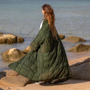Side profile view of the VEDA Grand duster in Forest Moss Green. The image captures the architectural silhouette of the heavy cotton canvas fabric. The ultra-wide kimono sleeve hangs with a dramatic, structured drape, characteristic of the Lagenlook style. Shows the deep, functional side pockets and the floor-sweeping maxi length. The deep green earth tone evokes a natural, organic vibe.