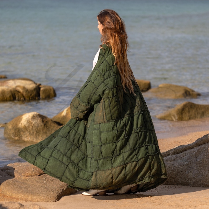 Side profile view of the VEDA Grand duster in Forest Moss Green. The image captures the architectural silhouette of the heavy cotton canvas fabric. The ultra-wide kimono sleeve hangs with a dramatic, structured drape, characteristic of the Lagenlook style. Shows the deep, functional side pockets and the floor-sweeping maxi length. The deep green earth tone evokes a natural, organic vibe.