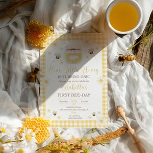 May include: A bee-themed invitation with the text "Our Little Honey is Turning One!" features a yellow gingham border and illustrations of bees and daisies. Honeycomb and a bowl of honey are also present, along with a wooden honey dipper.