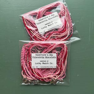 May include: Two clear plastic bags containing Valentine's Day friendship bracelets. Each bag has a label that reads "Valentine's Day Friendship Bracelets" and "Handmade by Lucky March Co." The bracelets are red, pink, and white.