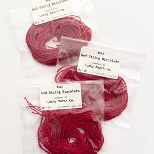 May include: Three clear plastic bags each containing a bundle of red string bracelets. Each bag has a white label with the text "BULK Red String Bracelets" and "HANDMADE BY Lucky March Co." The quantity is listed as 20.