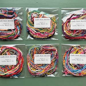 May include: Six clear plastic bags each containing a bundle of colorful friendship bracelets. Each bundle is a mix of red, blue, yellow, green, and other colors. Each bag has a white label with the text "Craft Room Seconds" and "Lucky March Co."