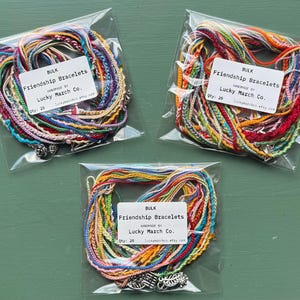 May include: Three clear plastic bags each containing a bundle of colorful friendship bracelets. Each bag has a white label that reads "BULK Friendship Bracelets" and "Lucky March Co." The bracelets are made of braided threads in various colors.