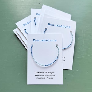 May include: Several white cards with blue text and a blue and green bracelet attached. The text on the cards reads "Beauxbatons" and "Academy of Magic Pyrenees Mountains Southern France". The background is a muted green.