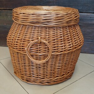 Puede incluir: Una cesta de mimbre tejida natural con tapa y asa circular. La cesta es de color marr&oacute;n y tiene un acabado natural.