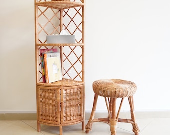 May include: A wicker corner shelf unit with three shelves and a cabinet with two doors. The shelf unit is filled with books, a tissue box, and a small wicker basket. A wicker stool is in front of the shelf unit.