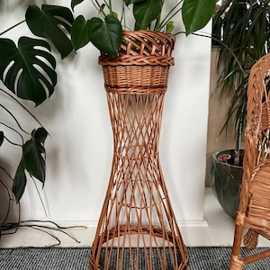 Op de afbeelding: Een hoge, geweven plantenstandaard van riet met een mandvormige bovenkant, met een weelderige groene plant. De standaard heeft een uitlopende basis en een roosterachtig ontwerp, met een natuurlijke bruine kleur. Een rieten stoel is gedeeltelijk zichtbaar.