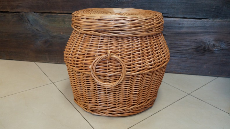 Puede incluir: Una cesta de mimbre tejida natural con tapa y asa circular. La cesta es de color marr&oacute;n y tiene un acabado natural.