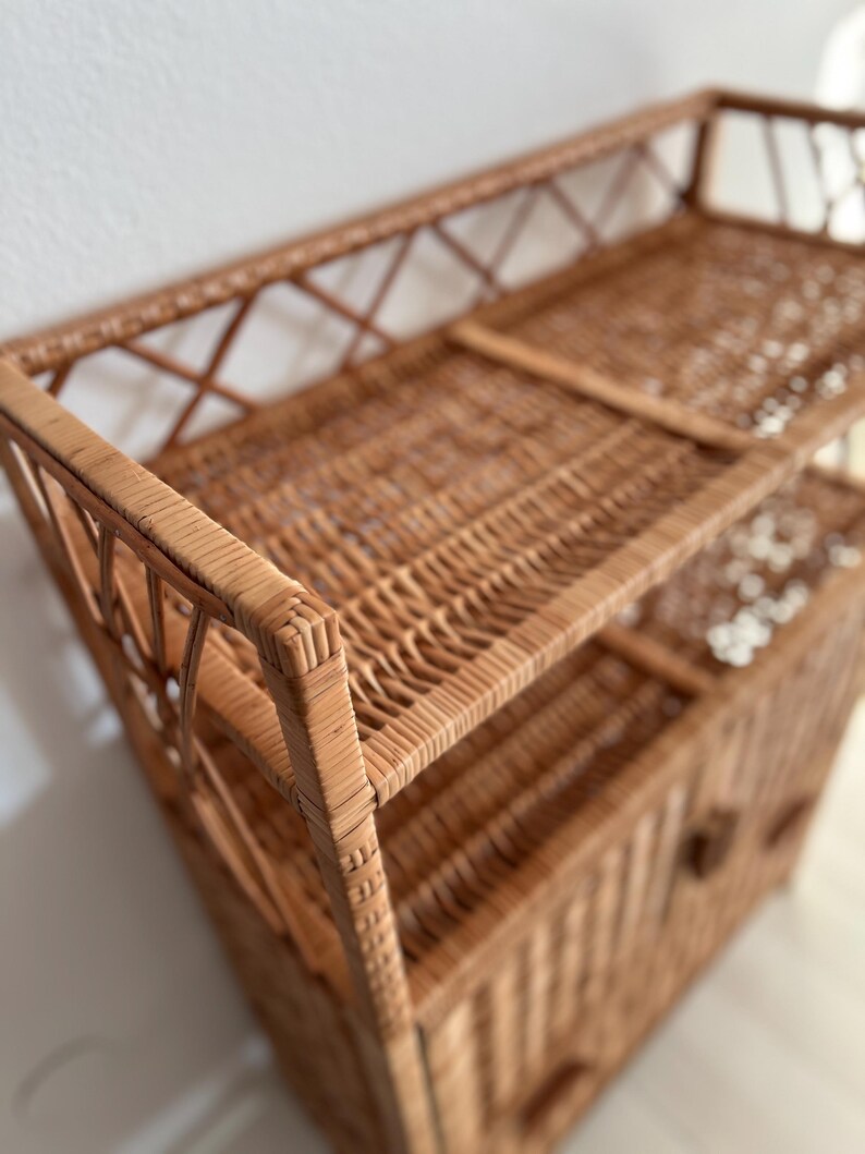 May include: A natural woven wicker shelf unit with two open shelves and a cabinet with a door. The cabinet has a small knob handle.
