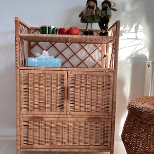 May include: A wicker cabinet with two doors and two shelves. The cabinet has a woven design and is made of natural materials. There are two apples and a green container on the top shelf. Two small dolls are on the top shelf.