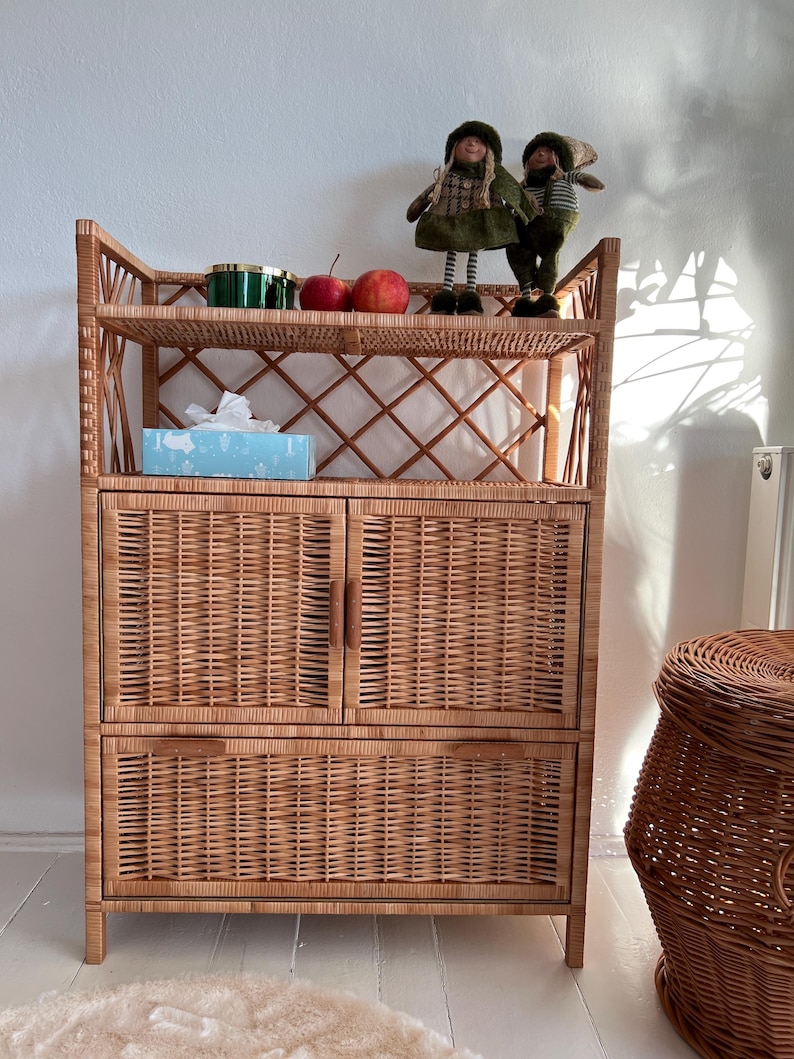 May include: A wicker cabinet with two doors and two shelves. The cabinet has a woven design and is made of natural materials. There are two apples and a green container on the top shelf. Two small dolls are on the top shelf.