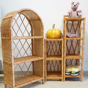 May include: Three wicker shelves, one with an arched top, are arranged side-by-side. The shelves are made of natural woven wicker and have a light brown color. A small, yellow pumpkin sits on the middle shelf of the smaller shelf unit. A pink crocheted teddy bear sits on the top shelf of the smaller shelf unit. A pair of fuzzy slippers are on the bottom shelf of the smaller shelf unit.