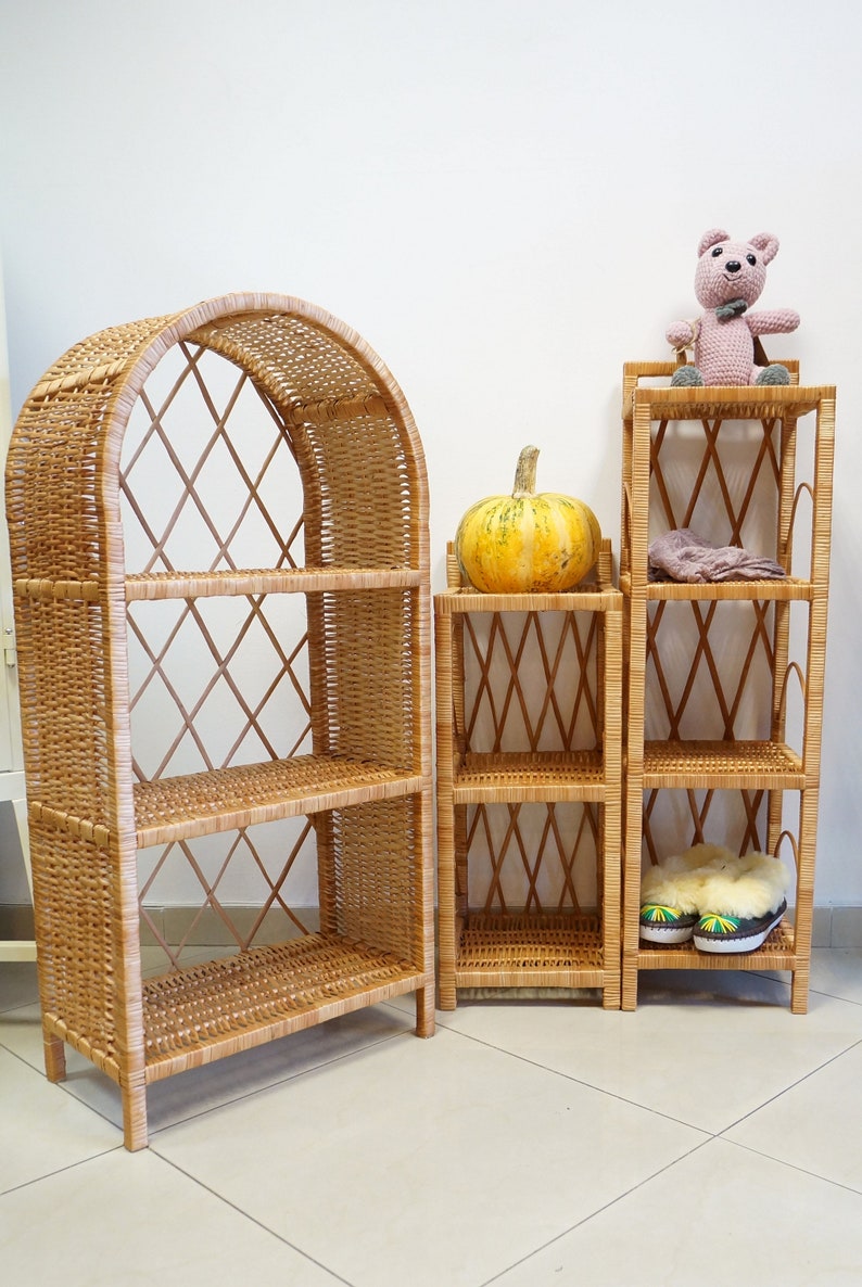 May include: Three wicker shelves, one with an arched top, are arranged side-by-side. The shelves are made of natural woven wicker and have a light brown color. A small, yellow pumpkin sits on the middle shelf of the smaller shelf unit. A pink crocheted teddy bear sits on the top shelf of the smaller shelf unit. A pair of fuzzy slippers are on the bottom shelf of the smaller shelf unit.