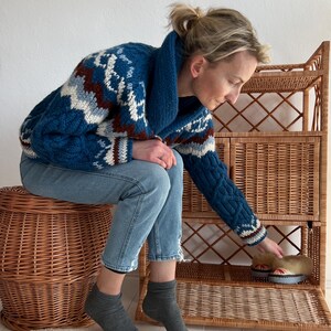 Puede incluir: Una mujer con jeans azules y un su&eacute;ter con estampado azul, blanco y marr&oacute;n se sienta en un taburete de mimbre y alcanza una estanter&iacute;a de mimbre. La estanter&iacute;a tiene una puerta abierta, que revela un par de zapatillas marrones y blancas.
