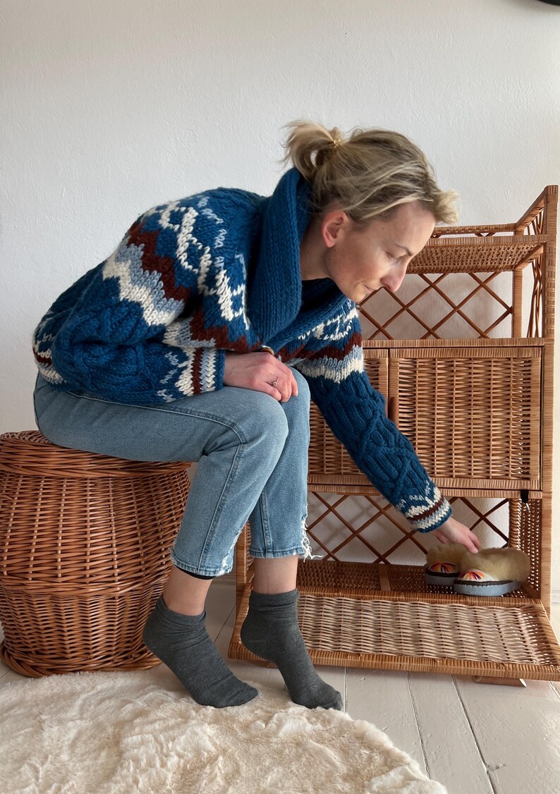 Puede incluir: Una mujer con jeans azules y un su&eacute;ter con estampado azul, blanco y marr&oacute;n se sienta en un taburete de mimbre y alcanza una estanter&iacute;a de mimbre. La estanter&iacute;a tiene una puerta abierta, que revela un par de zapatillas marrones y blancas.