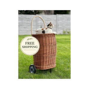 Op de afbeelding: Een bruine rieten dierenmand met een gebogen handvat en wielen, op groen gras. Een kat met zwarte en bruine vlekken zit in de mand. De mand heeft een ronde sticker met de tekst "FREE SHIPPING".
