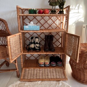 May include: A wicker shelf unit with four shelves and two doors. The shelves are filled with shoes, a box of tissues, and decorative items. The shelf unit is standing on a white rug.