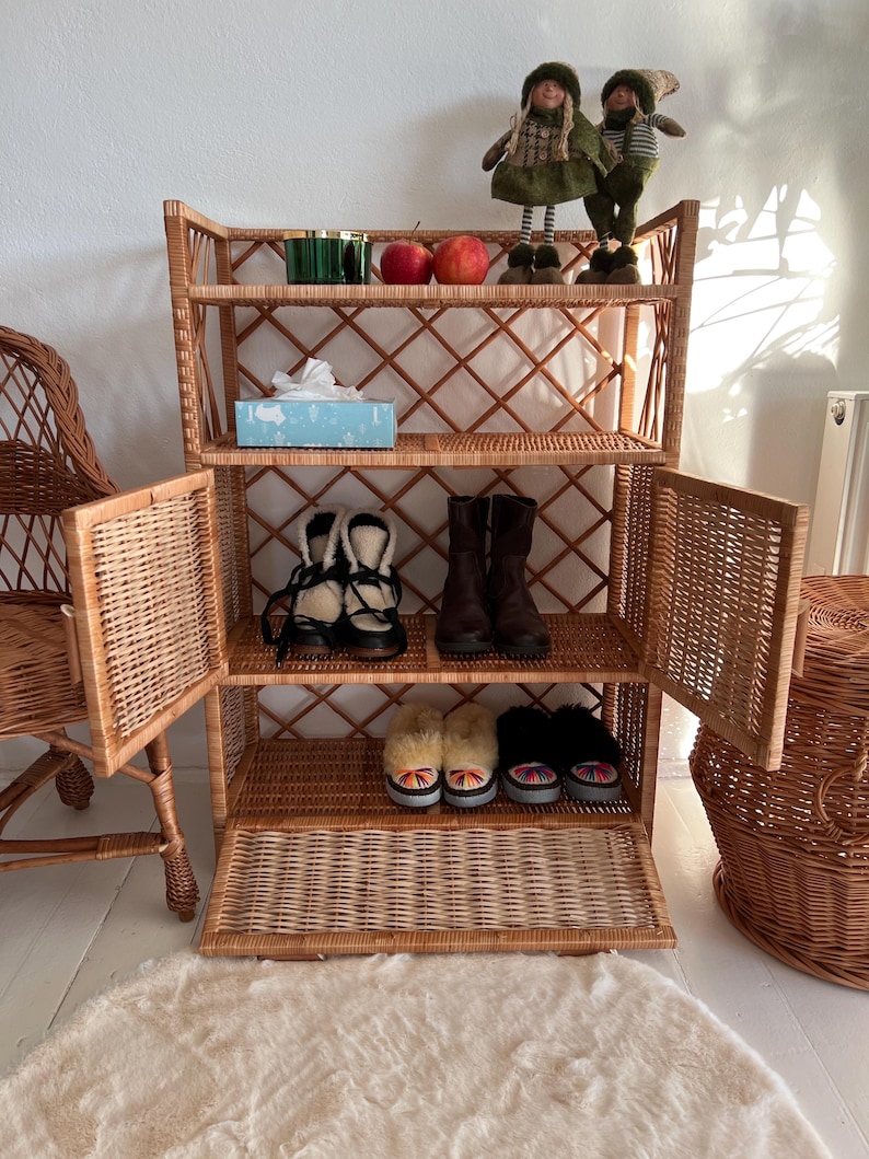 May include: A wicker shelf unit with four shelves and two doors. The shelves are filled with shoes, a box of tissues, and decorative items. The shelf unit is standing on a white rug.