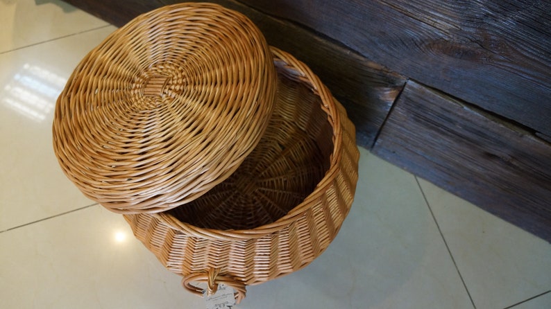 Puede incluir: Una cesta redonda de mimbre con tapa. La cesta est&aacute; hecha de mimbre tejido natural y tiene un color marr&oacute;n claro. La tapa est&aacute; ligeramente entreabierta, revelando el interior de la cesta.