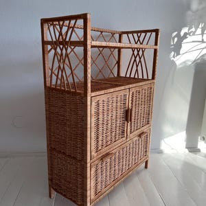 May include: A light brown wicker cabinet with two doors and two shelves. The cabinet has a woven design and is standing on a white wooden floor.