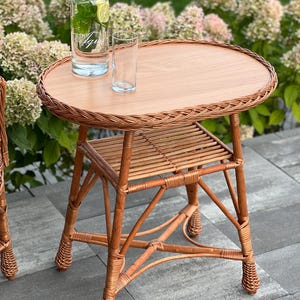 Peut inclure: Une table d'appoint ronde en osier brun clair avec un plateau en bois. La table a une étagère inférieure et est associée à une bouteille d'eau en verre transparent et un verre à boire. La table est à l'extérieur, entourée de verdure.