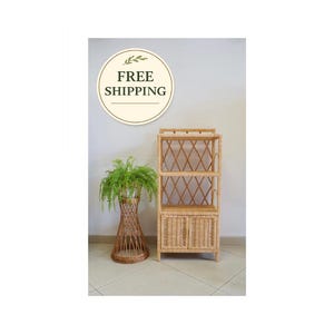 Könnte beinhalten: Ein natürliches Bücherregal aus Rattan mit drei Regalen und einem Schrank. Die Regale haben ein Rautenmuster und die Schranktüren sind gewebt. Neben dem Bücherregal steht eine hohe, gewebte Vase mit einem Farn. Das Bild enthält eine "FREE SHIPPING" Grafik.