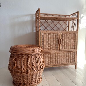Puede incluir: Un armario de almacenamiento de mimbre con dos puertas y un estante, junto a una cesta de almacenamiento de mimbre con asa.